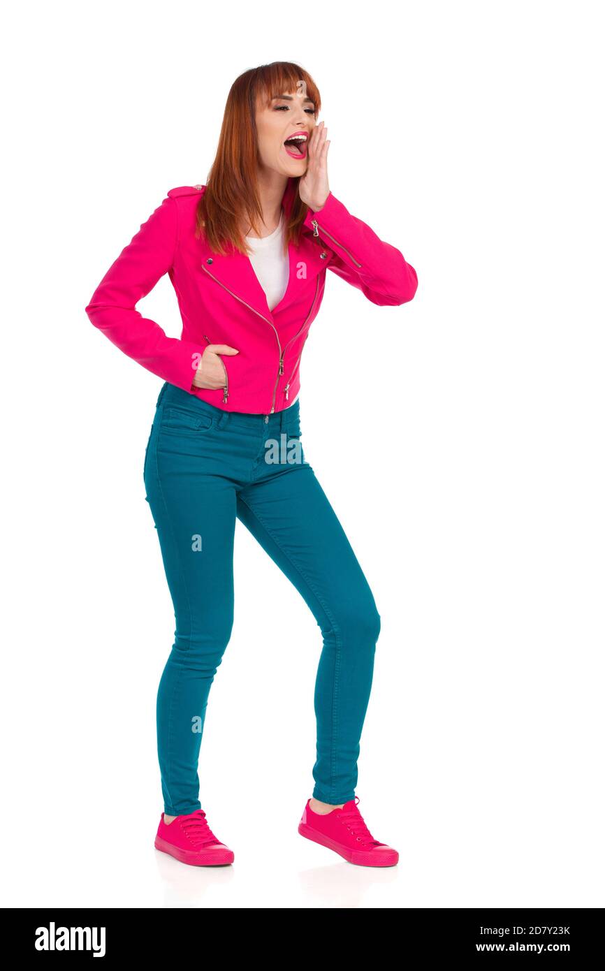 Aufgeregte junge Frau in rosa Jacke, grünen Jeans und Turnschuhen steht, Hand am Kinn haltend und schreiend. Seitenansicht. Studioaufnahme in voller Länge, Isolat Stockfoto