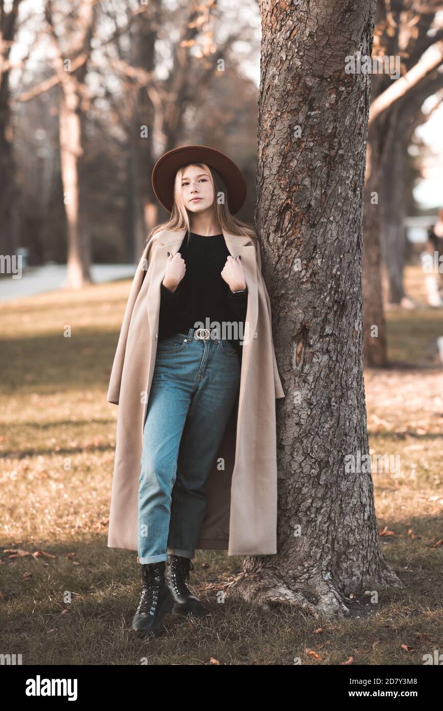 Stilvolle Teenager-Mädchen 14-15 Jahre alt trägt trendigen Wintermantel und Hut posiert im Park lehnen sich auf Baum im Freien. Blick auf die Kamera. Teenagerhood. Stockfoto