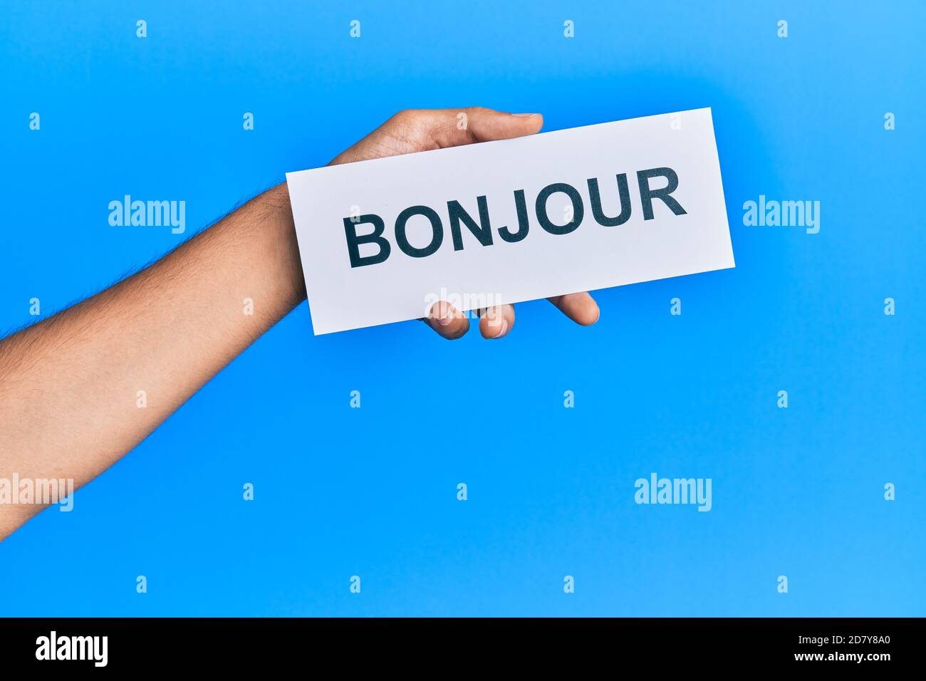 Hand von hispanischen Mann hält französisch Gruß bonjour Wort Papier über isolierten blauen Hintergrund. Stockfoto