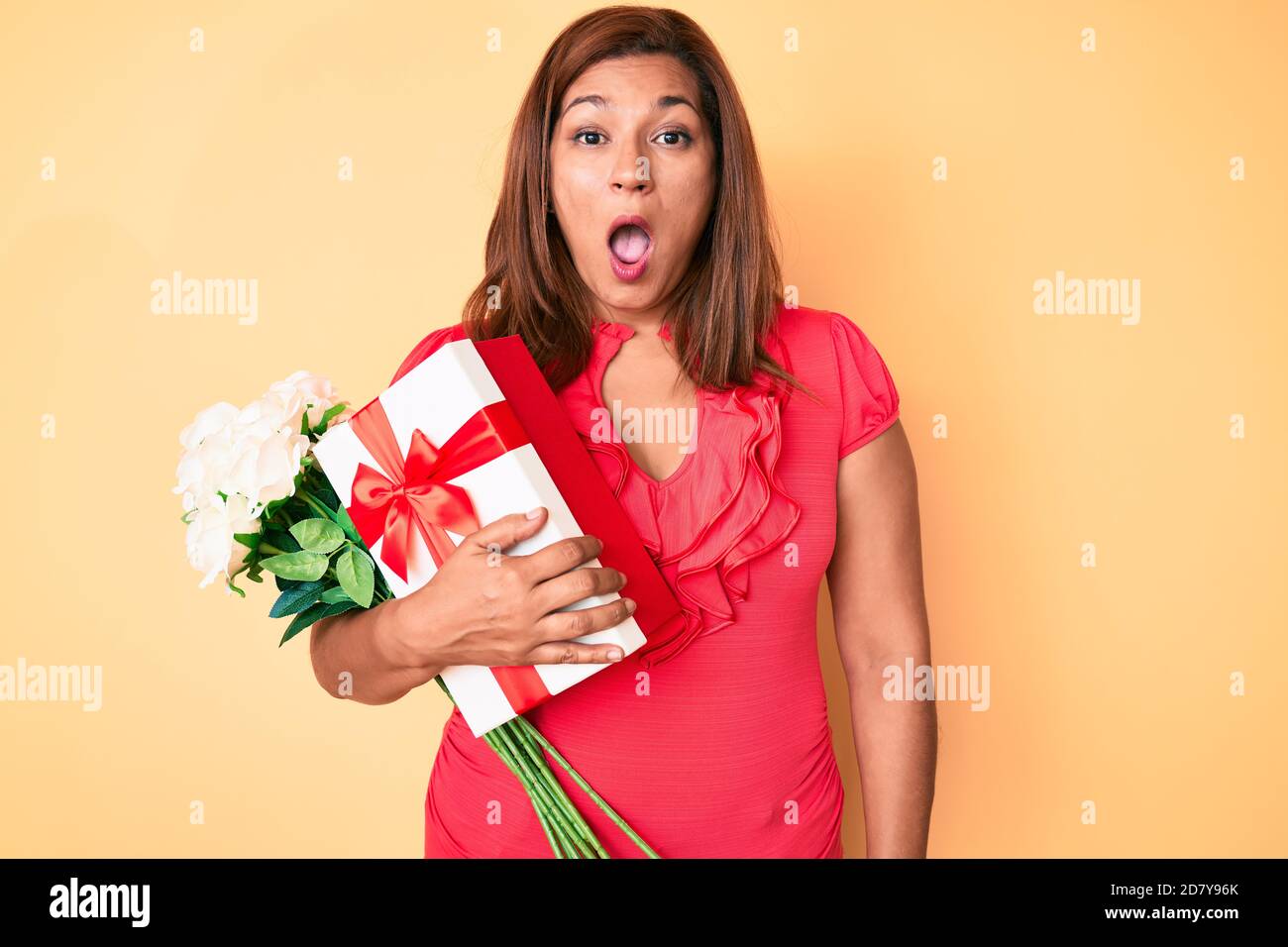 Mittleres Alter Brünette Frau hält Jahrestag Geschenk und Blumenstrauß verängstigt und erstaunt mit offenem Mund für Überraschung, Unglauben Gesicht Stockfoto