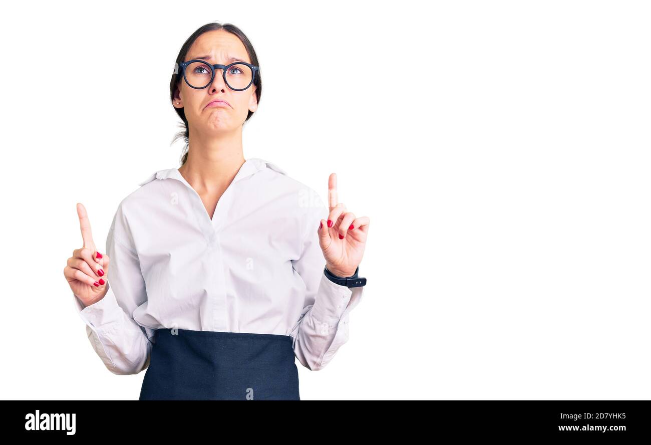 Schöne Brünette junge Frau trägt professionelle Kellnerin Schürze zeigt nach oben traurig und aufgeregt, zeigt Richtung mit den Fingern, unglücklich und d Stockfoto