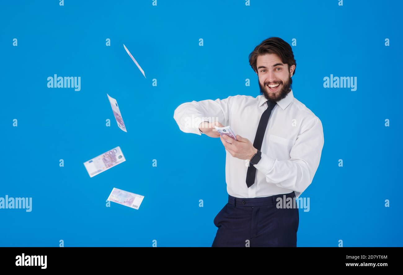 Ein junger fröhlicher bärtiger Geschäftsmann in einem weißen Hemd und schwarzer Krawatte auf blauem Hintergrund spritzt Geld. Verrückter Kerl wirft Geldscheine. Studio Stockfoto