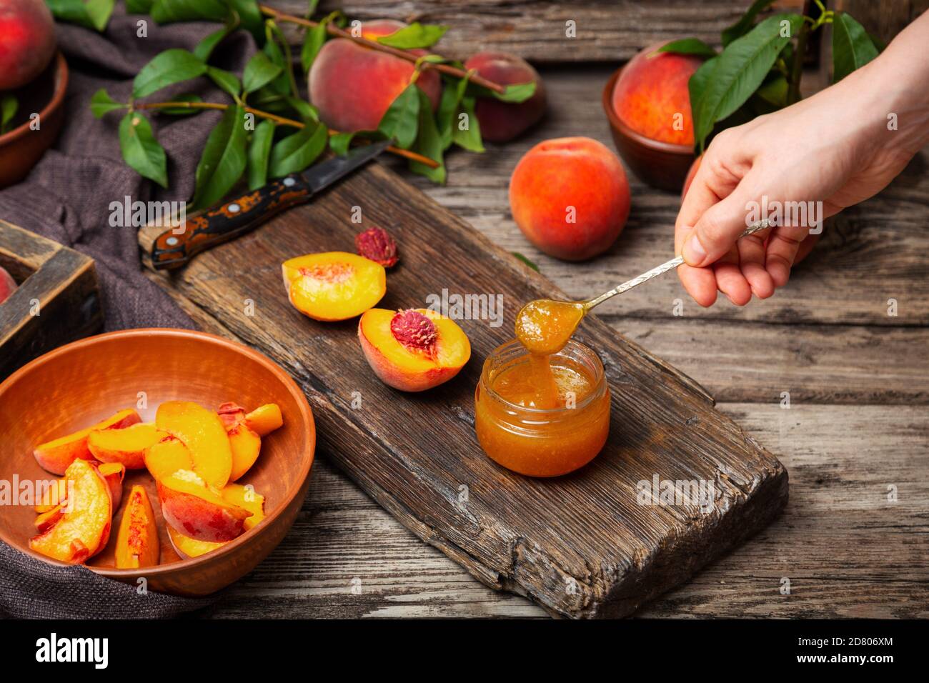 Pfirsich Marmelade Löffel in weiblicher Hand. Konservenglas mit Pfirsichkeilen und ganzen Früchten. Pfirsichmarmelade auf Holztisch mit Zutaten. Prozess von Stockfoto