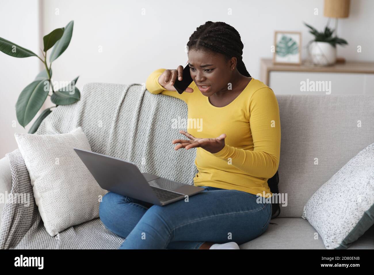 Frustrierte schwarze Dame mit Laptop und im Gespräch auf Handy an Zu Hause Stockfoto