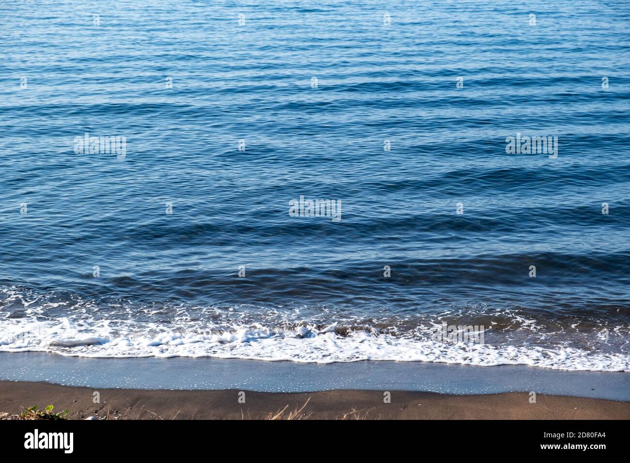 Sommerferienkonzept. Blauer ruhiger Meereshintergrund trifft auf den Sandstrand. Schaum und Reflexionen auf dem Wasser, am leeren Ufer früh am Morgen. Stockfoto