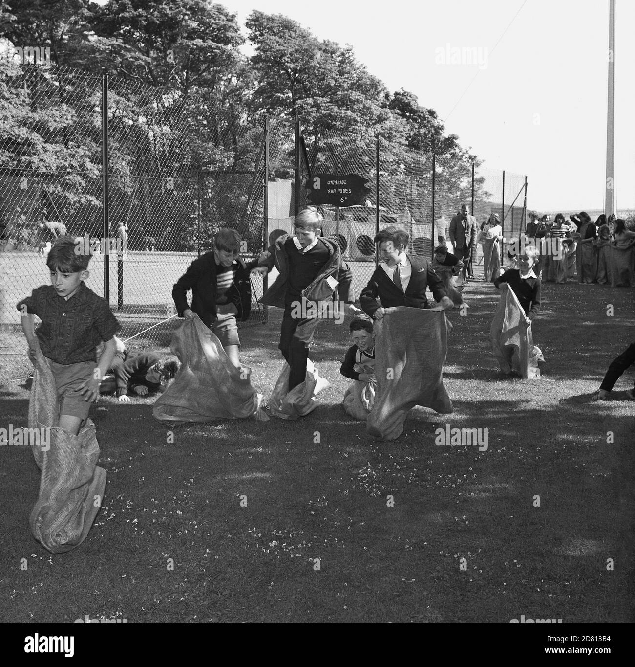 60er Jahre, historisch, Schüler, die an einem Sackrennen auf einer Wiese an einer Schule oder einem Dorffest teilnehmen, Fife, Schottland, mit einem S-Schild, das sagt, 'Stoneage' Kar Rides kostet 6d. Stockfoto