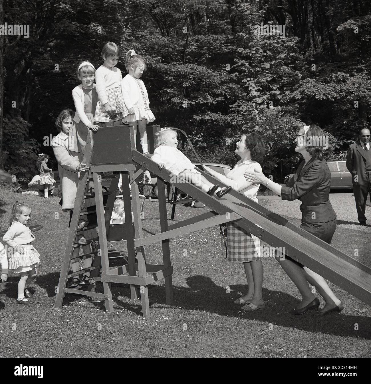 1960er Jahre, draußen auf einem Feld in einem Park, warten kleine Mädchen auf ihre Reihe, um eine hölzerne Rutsche zu gehen, mit ihren Müttern zur Hand, um sie zu fangen, Fife, Schottland. Stockfoto