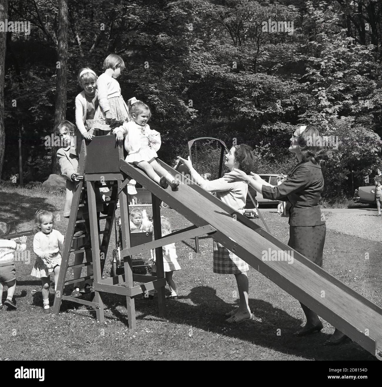 1960er Jahre, draußen auf einem Feld in einem Park, warten kleine Mädchen auf ihre Reihe, um eine hölzerne Rutsche zu gehen, mit ihren Müttern zur Hand, um sie zu fangen, Fife, Schottland. Stockfoto