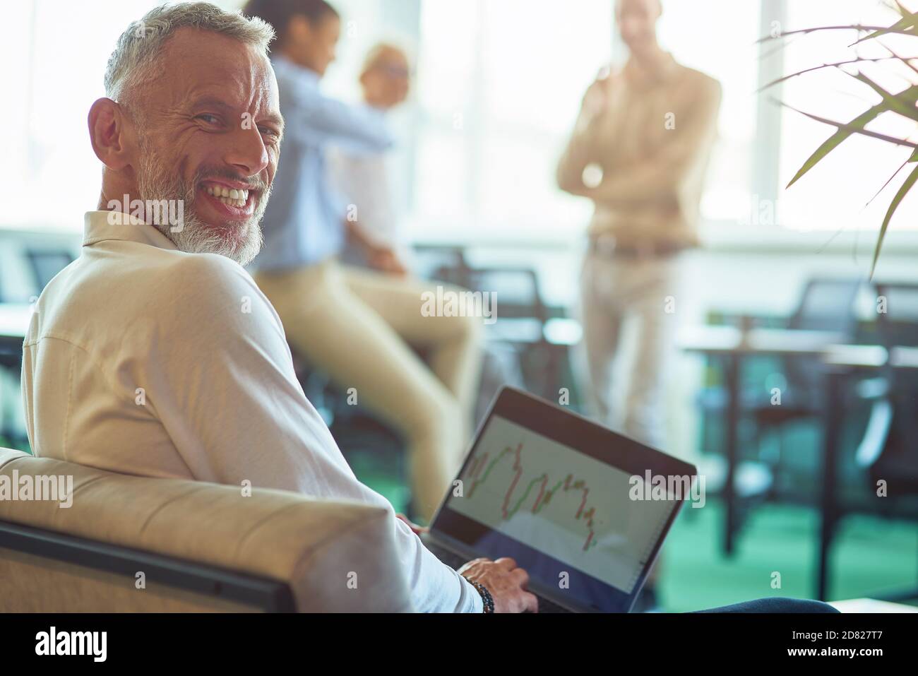 Junger, fröhlicher, reifer Geschäftsmann sitzt im modernen Büro, mit Laptop und lächelt vor der Kamera, während seine Kollegen im Hintergrund sprechen Stockfoto