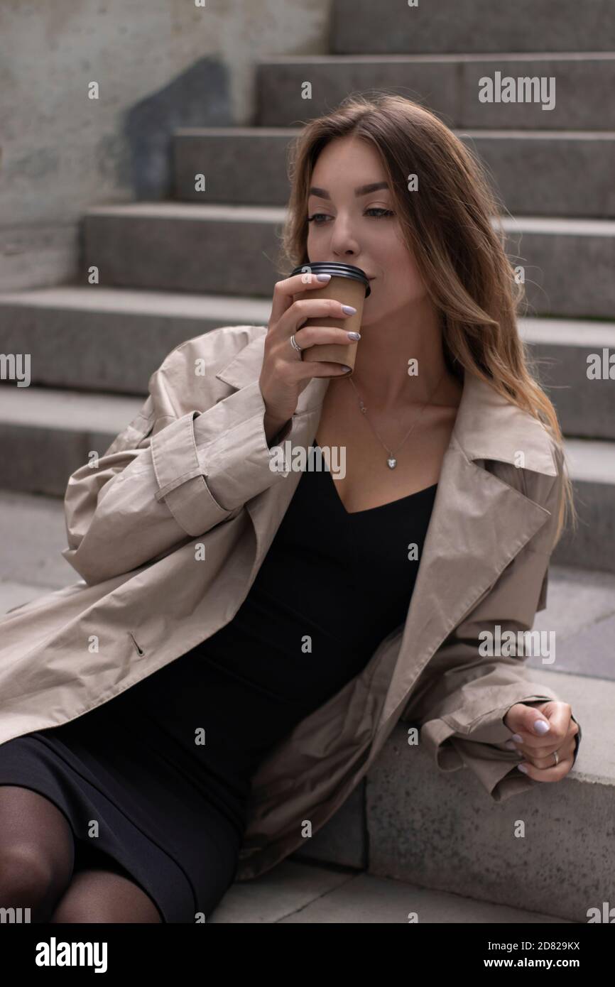 Attraktive schlanke junge Brünette in modischen Graben und schwarzen Kleid sitzt auf grauen Betontreppe mit einem braunen Papiertasse Kaffee. Stockfoto