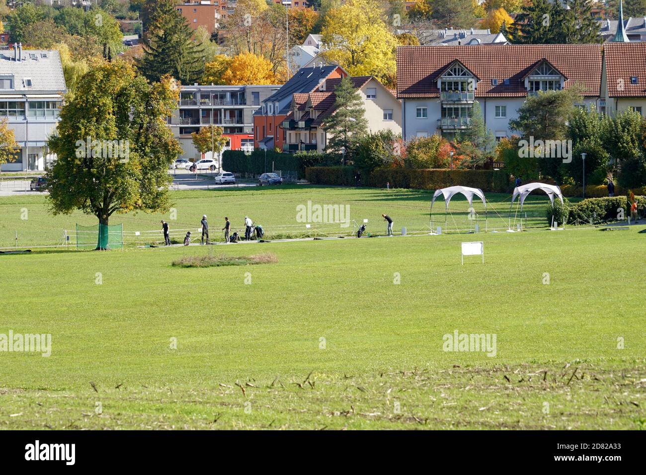 Golfclub-Mitglieder im Club Urdorf an einem sonnigen Herbstsamstagnachmittag spielen und üben mit Dorfzentrum im Hintergrund. Stockfoto