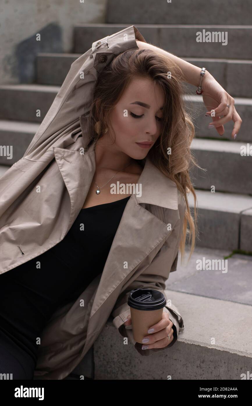 Attraktive schlanke junge Brünette in modischen Graben und schwarzen Kleid sitzt auf grauen Betontreppe mit einem braunen Papiertasse Kaffee. Stockfoto