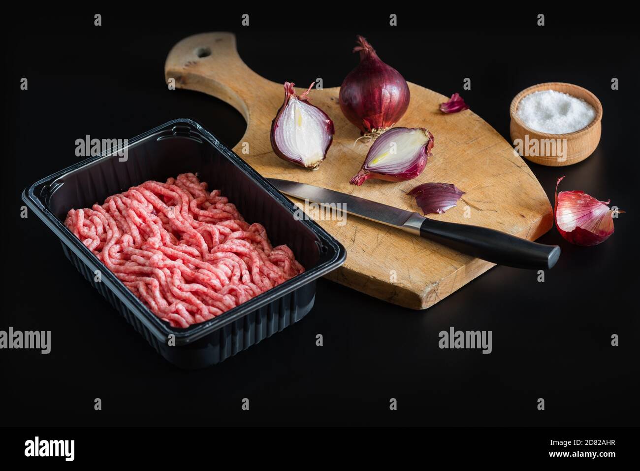 Hackfleisch ist ein Halbzeug in gewerblichen Verpackungen. Ein Messer und rote Zwiebeln liegen auf einem hölzernen Schneidbrett. Salzstreuer mit Salz. Schwarz-Bac Stockfoto