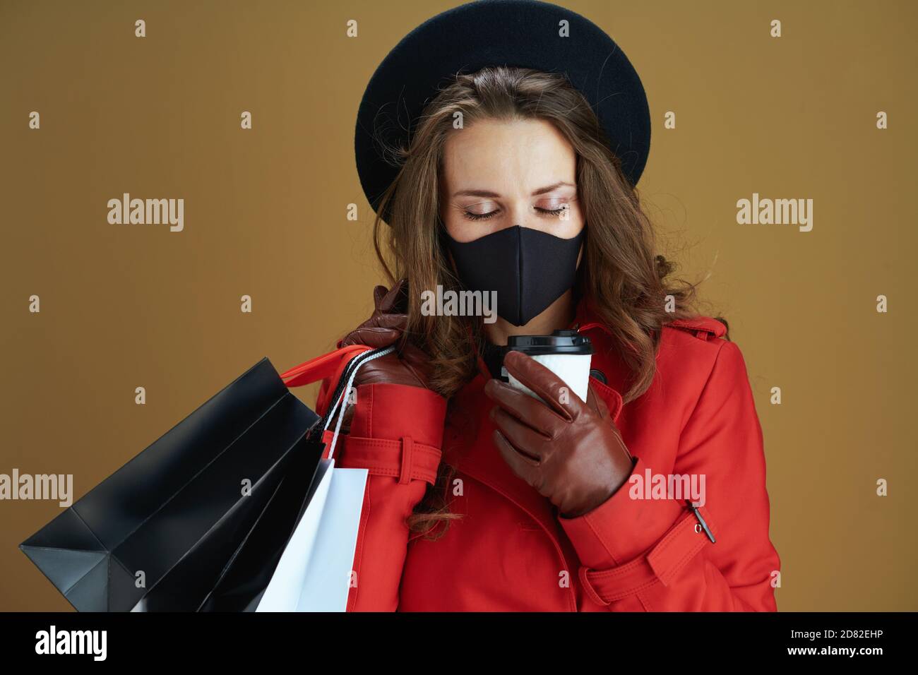 Leben während Coronavirus Pandemie. Stilvolle Hausfrau in rotem Mantel mit schwarzer Maske, Tasse und Papier Einkaufstaschen vor Bronze Hintergrund. Stockfoto