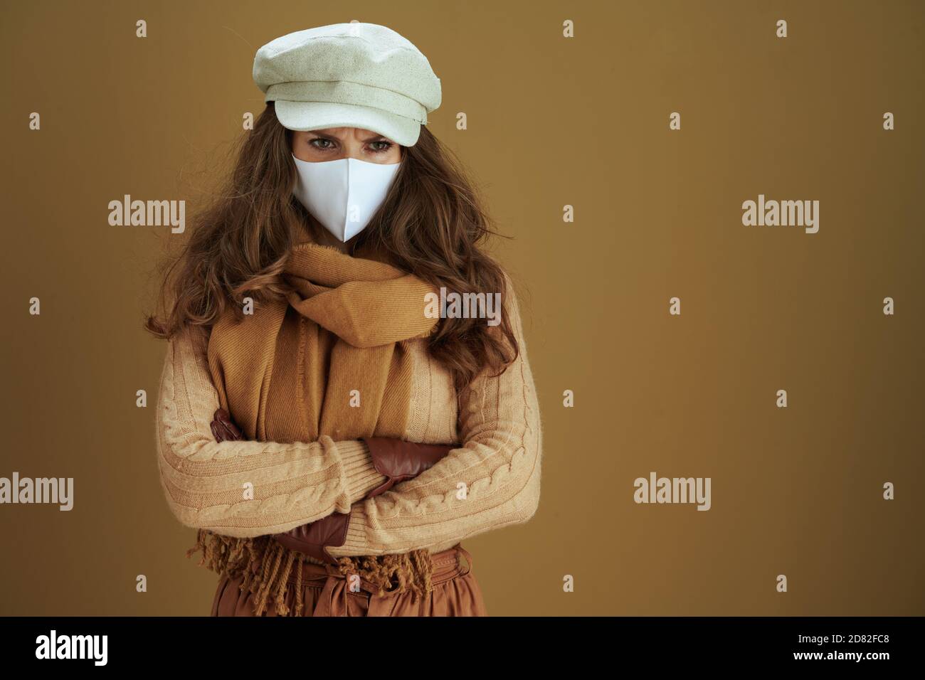 Leben während der Coronavirus-Pandemie. Trendige Hausfrau in Pullover mit weißen medizinischen Maske auf Bronze Hintergrund. Stockfoto