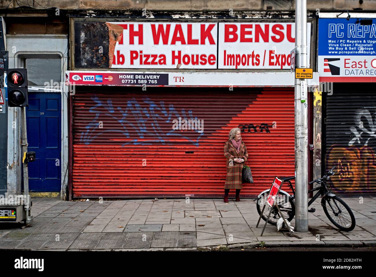 Eine elegant gekleidete reife Frau wartet vor einem heruntergekommenen und geputzten Pizzaladen auf Leith Walk, Edinburgh, Schottland, Großbritannien. Stockfoto