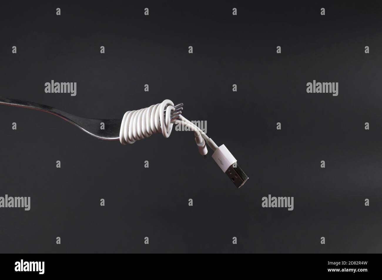 Ladekabel des Mobiltelefons, das wie Spaghetti um die Gabel gewickelt ist. Dunkelgrauer Hintergrund mit Gabel und Netzkabel vorne. Konzeptionelle Fotografie. Stockfoto