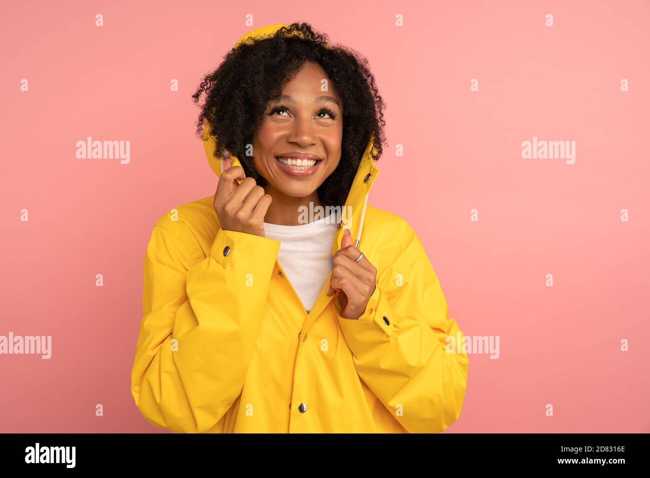 Lächelnde dunkelhäutige lockige Frau in gelber Regenjacke mit Kapuze hat gute Laune, Blick auf Regen und leeren Raum für Werbung, Werbeinhalte, Stockfoto