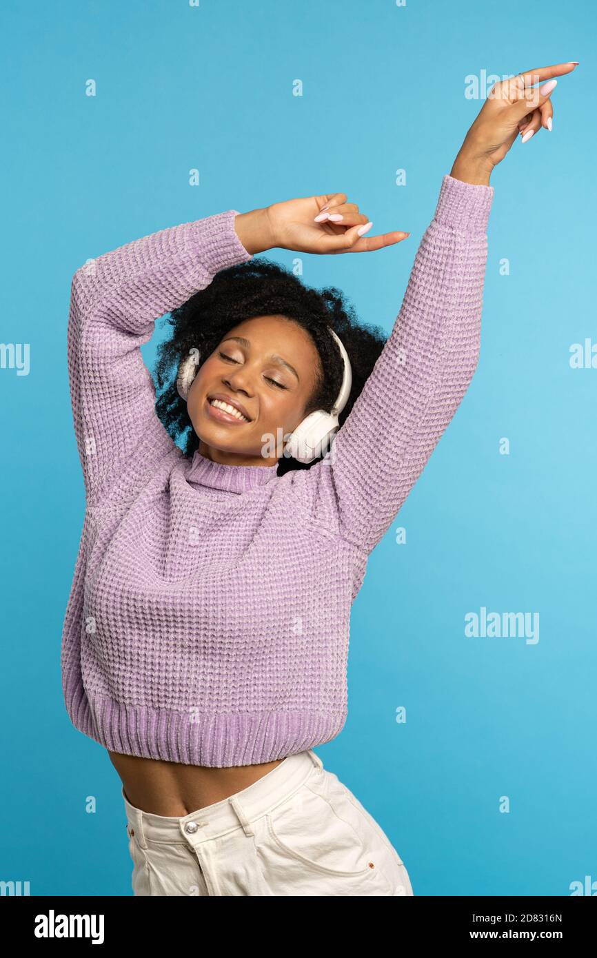 Energetisch Biracial lockig Frau tanzen, Lächeln breit, trägt kabellose Kopfhörer, hat gute Laune, hält die Hände in der Luft angehoben, isoliert auf blauem Hintergrund Stockfoto