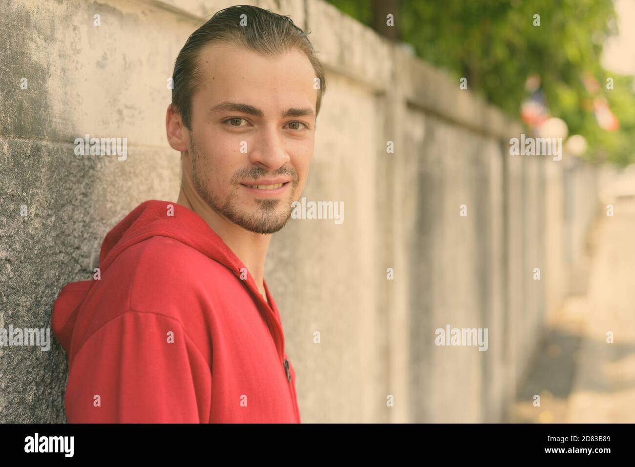 Junge glücklich bärtiger Mann lächelnd, während das Tragen der roten Hoodie und lehnte sich auf alte Betonwand in den Straßen im Außenbereich Stockfoto