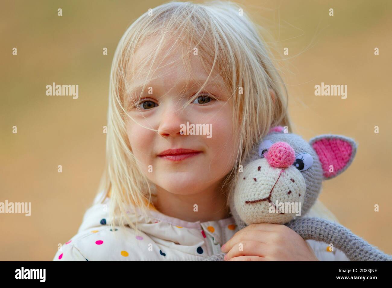 Nahaufnahme Porträt der schönen blonden Kleinkind Mädchen Umarmung ihrer Lieblings-handgemachte Spielzeug (gestrickte Katze) im Freien. Windiger Tag, unordentliche Haare im Gesicht. Stockfoto