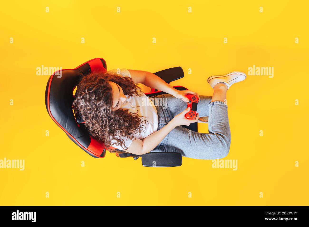 Frau, die hinter einem Spielstuhl in ihren Händen sitzt und roten Gamepad auf gelbem Hintergrund hält, Blick von oben Stockfoto