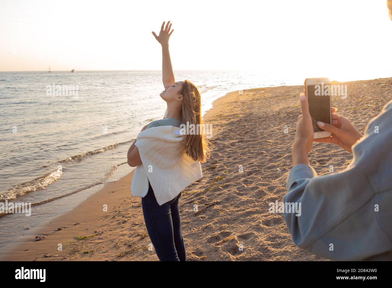 Junge Frau macht Fotos von ihrer Freundin am Strand gegen den Sonnenuntergang, Foto für soziale Netzwerke. Stockfoto