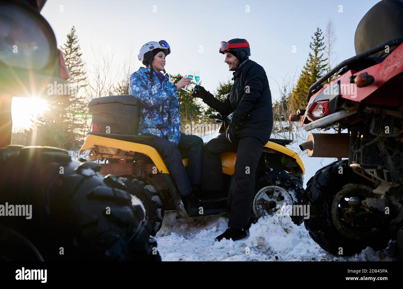 Junges Paar in Winterskianzügen und Helmen, auf gelbem ATV sitzend, zwei Gläser mit blauem Champagner haltend, die sich gegenseitig anschauten. Sonniger Wintertag. Riesige Räder anderer ATV stehen im Vordergrund Stockfoto