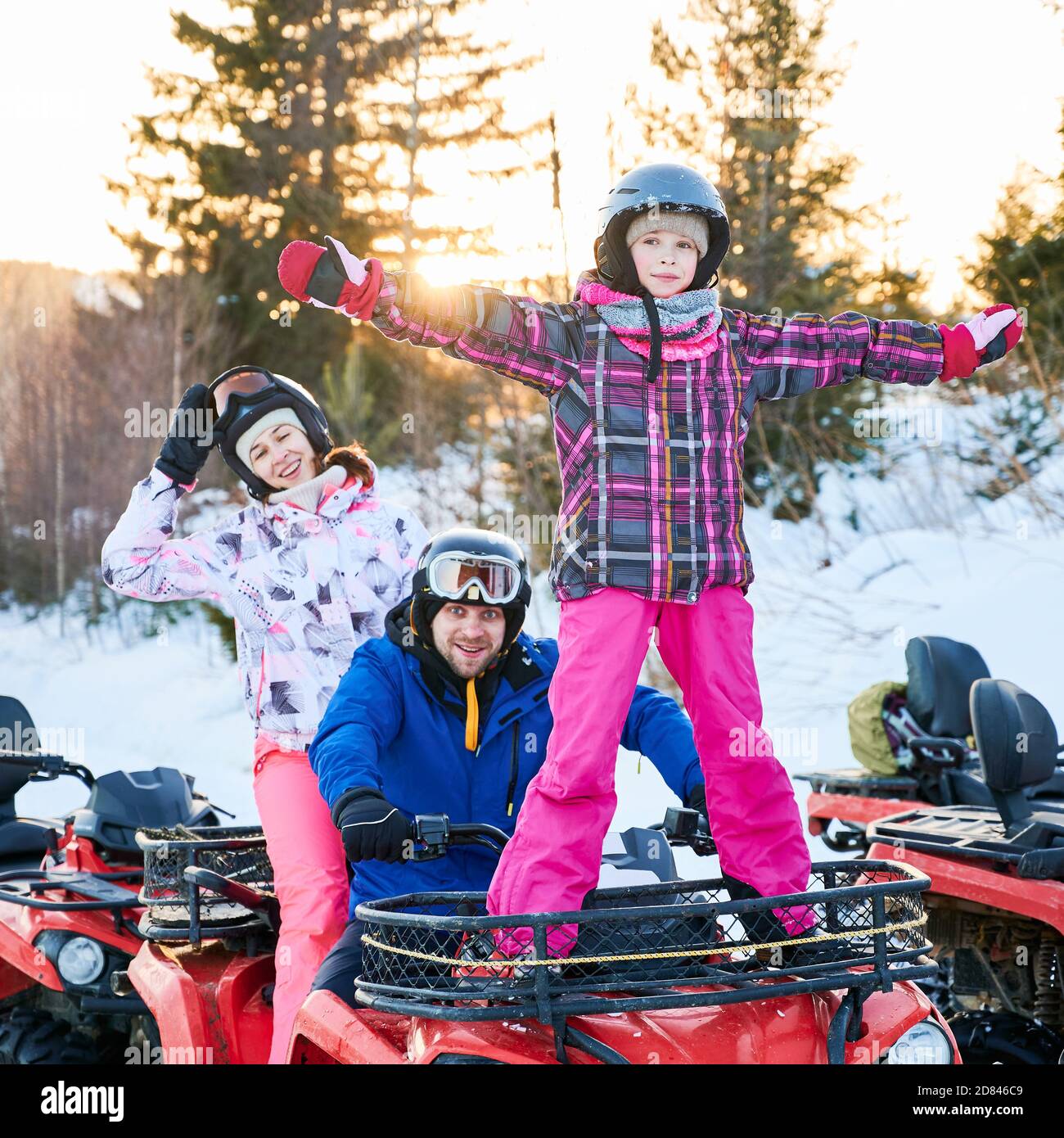 Junge Familie in Helmen lächelt zur Kamera, während sie auf Geländewagen mit niedlichen Tochter sitzt. Liebenswert kleines Mädchen in Winterjacke genießen Quad Radfahren mit Eltern. Konzept aktive Freizeit. Stockfoto