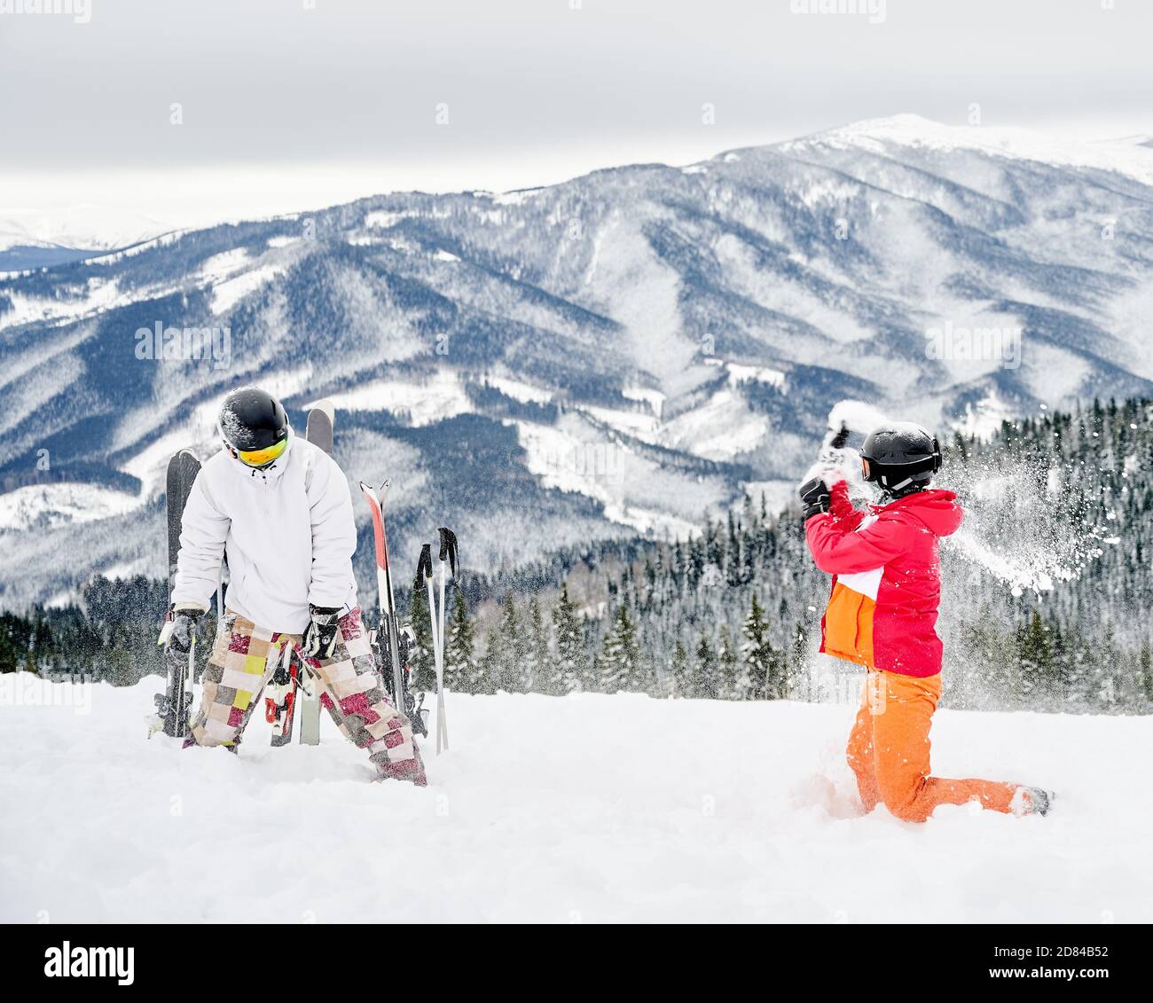 Frau und Mann Skifahrer in Skianzügen und Helmen spielen mit Pulverschnee. Paar Spaß auf verschneiten Hügel mit schönen Bergen im Hintergrund. Konzept des Skifahrens, aktive Freizeit und Beziehungen. Stockfoto