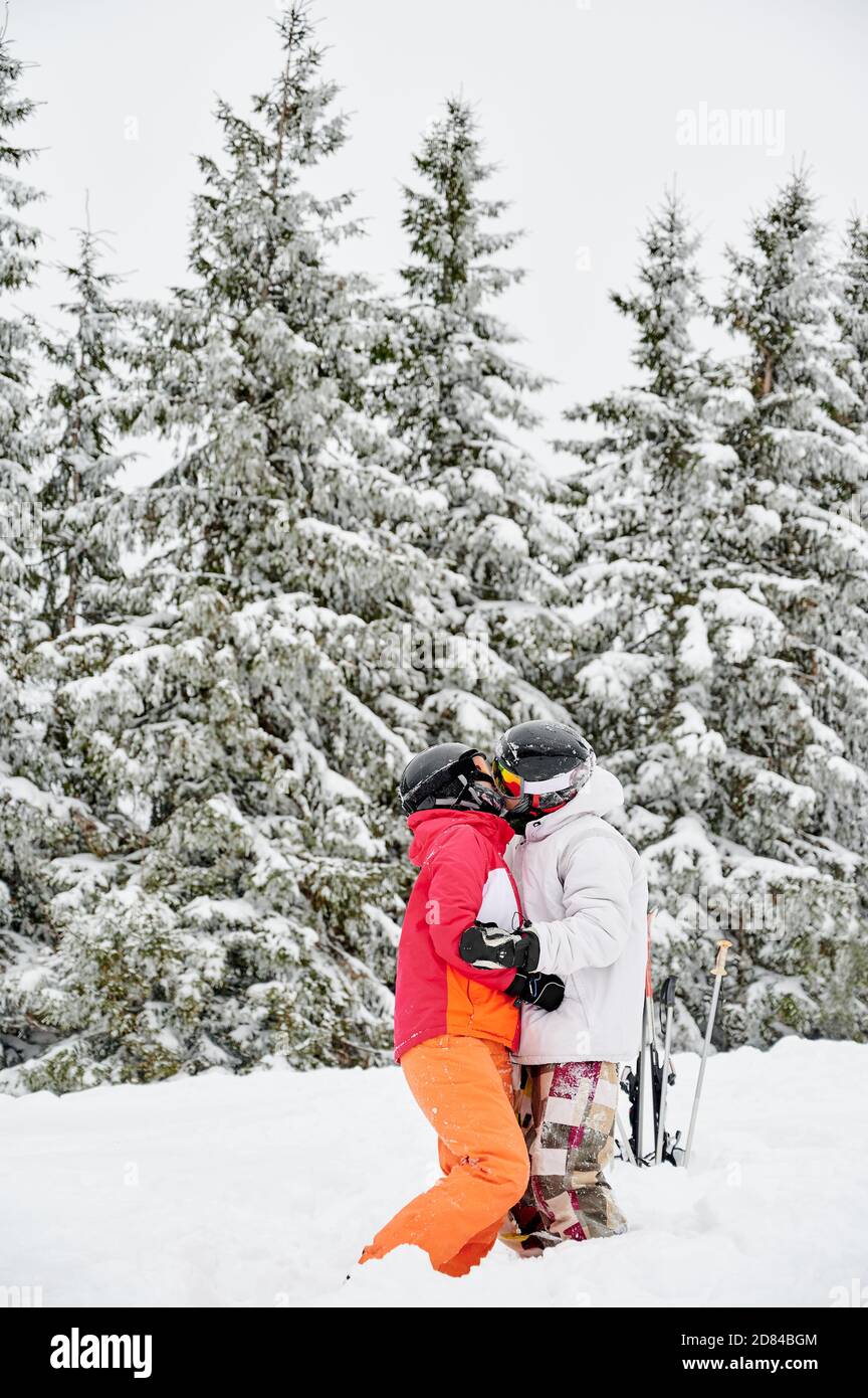 Vertikale Momentaufnahme von jungen paar Touristen küssen im Schnee gegen hypnotisierende schneebedeckten Fichtenwald auf Hintergrund. Lebendige bunte Anzüge auf weißem Schnee. Konzept der Romantik Stockfoto