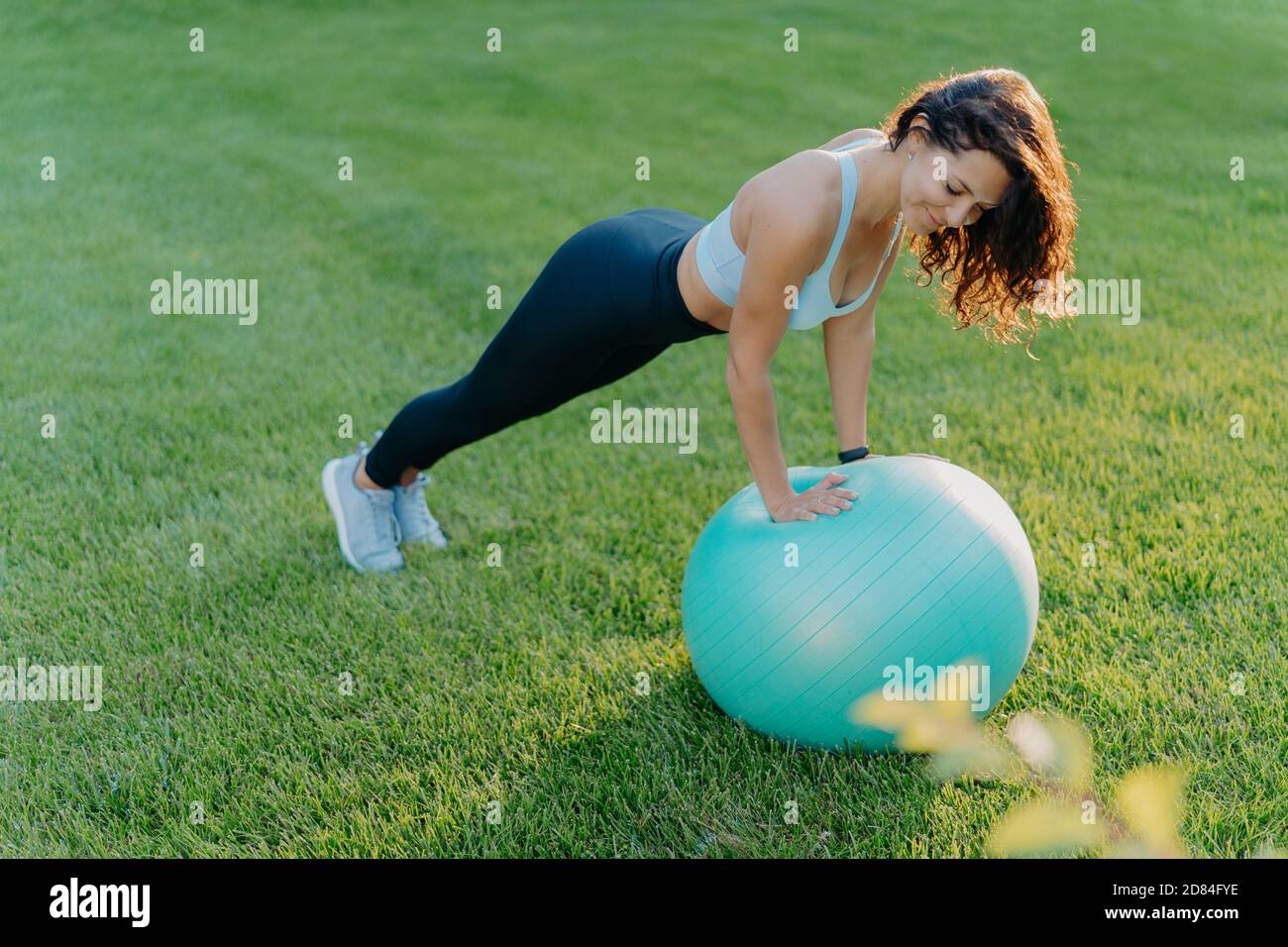 Sportliche Mädchen steht in Plank Pose auf Gymnastikball, in Sportkleidung gekleidet, macht körperliche Übungen auf grünem Rasen im Freien, hält fit und gesund. Peopl Stockfoto