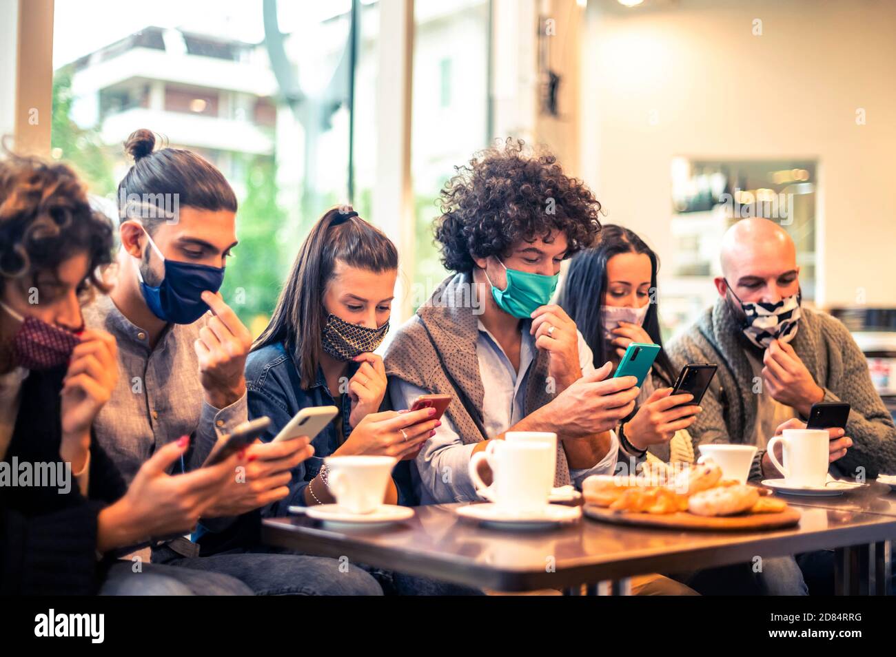 Multirassische Freunde mit Gesichtsmaske halten Smartphones sitzen am Tisch Cappuccino trinken im Café-Shop in der covid-19 Zeit - Leute, die Spaß haben zu toge Stockfoto