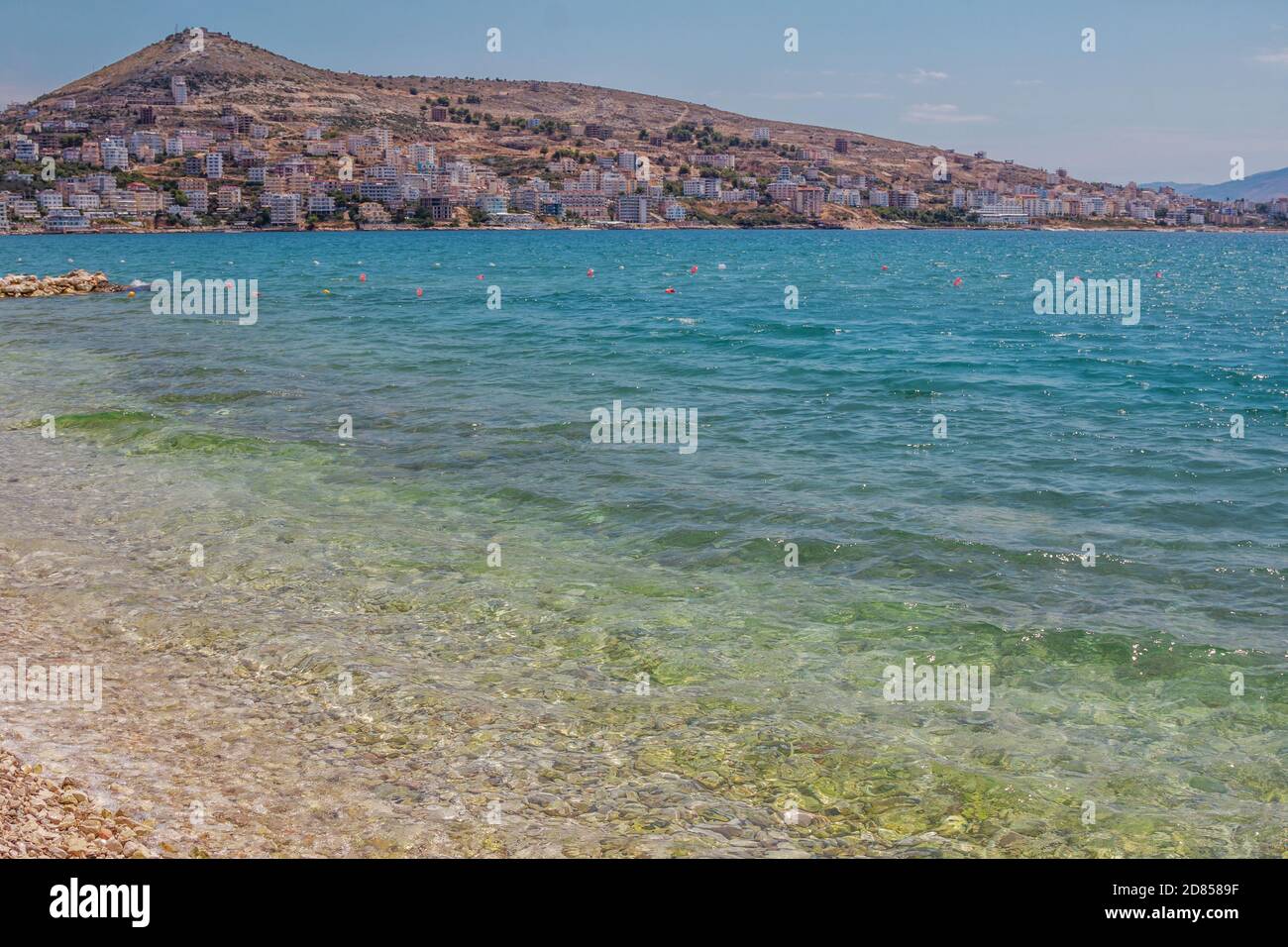 Blick auf das wunderschöne albanische Resort – Saranda. Landschaft mit klarem türkisfarbenem Meerwasser, Gebäuden auf einem Hügel und blauem Himmel. Stockfoto