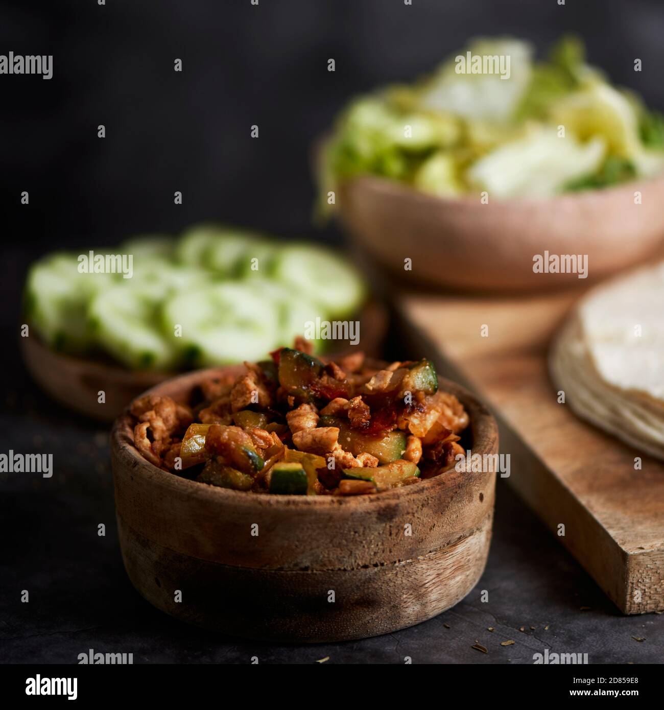 Eine Schüssel mit einem Huhn Fleisch und Gemüse Füllung auf einem Tisch, neben einem Teller mit einigen Scheiben Gurke, eine Schüssel mit Salat und einige Tortillas oder Stockfoto