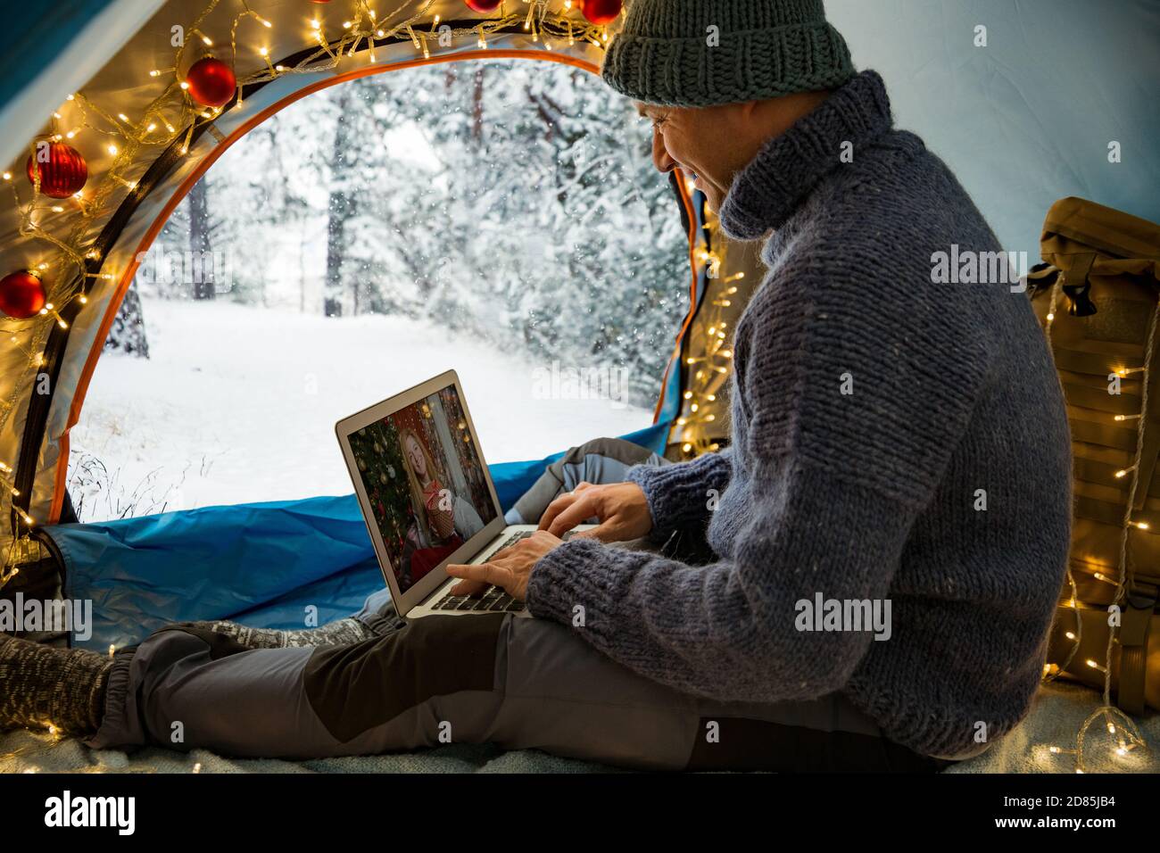 Mann sitzt im Zelt mit Weihnachtsbeleuchtung geschmückt, mit Laptop. Videoanruf. Skandinavische Landschaft, mit Schnee bedeckter Wald. Selbstisolierung Stockfoto