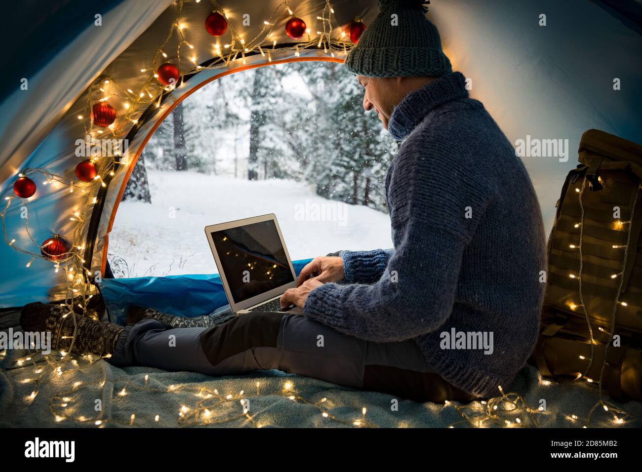 Mann sitzt im Zelt mit Weihnachtsbeleuchtung geschmückt, mit Laptop. Schöne Draufsicht auf skandinavische Landschaft, mit Schnee bedeckter Wald. Stockfoto