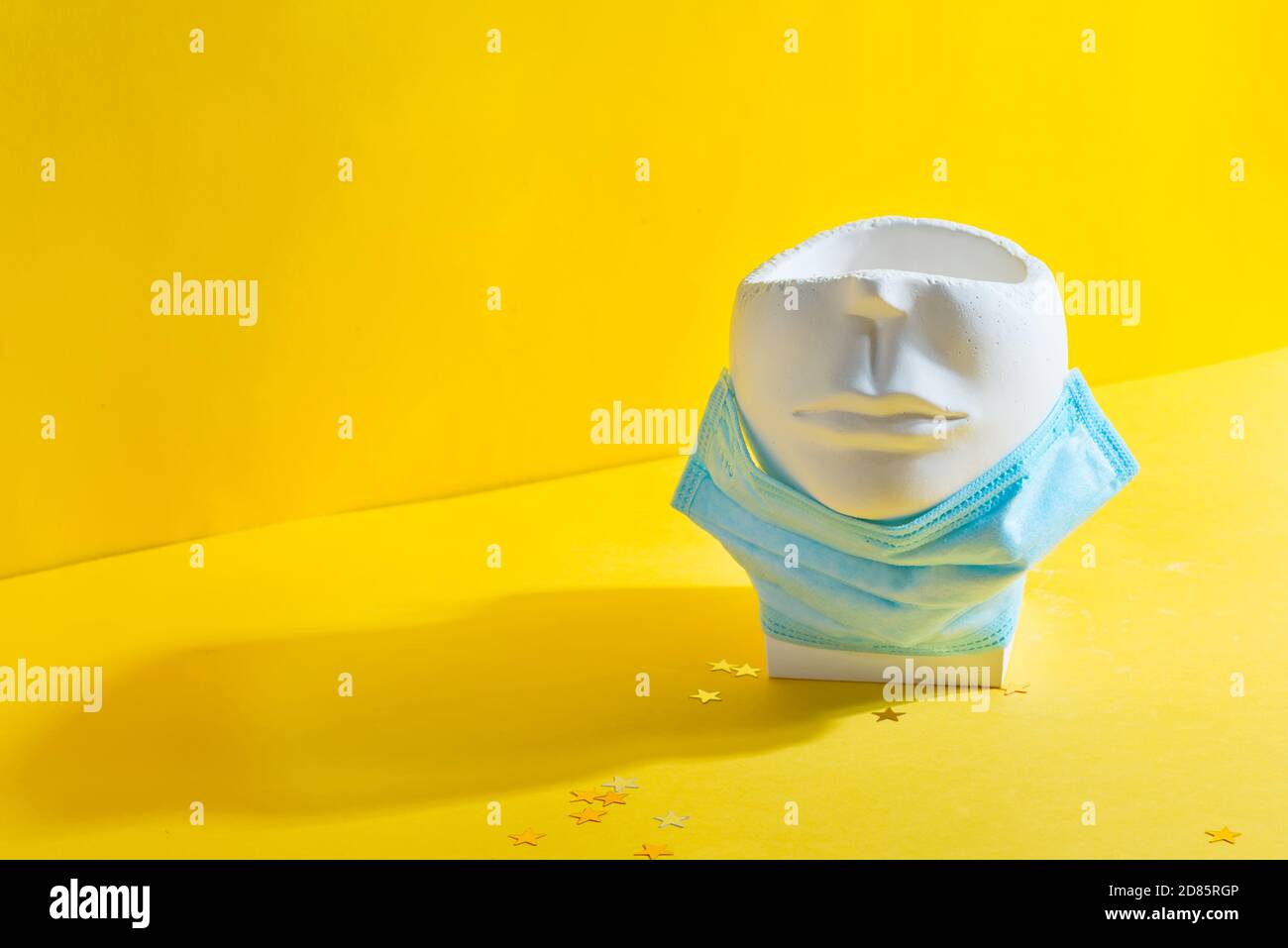 Gipskunsttopf in Form von menschlichem Gesicht mit schützender medizinischer Maske unter dem Kinn auf gelbem Hintergrund mit Schatten, Kopierraum. Stockfoto