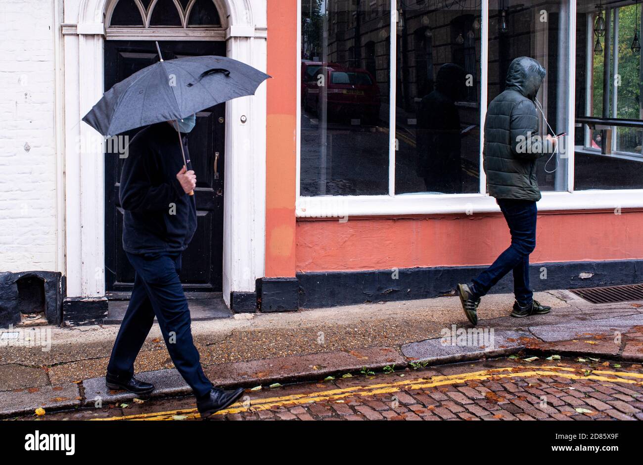 Mann mit Gesichtsmaske, der am Gebäude mit Regenschirm vorbeiläuft, Norwich, Norfolk, Großbritannien Stockfoto