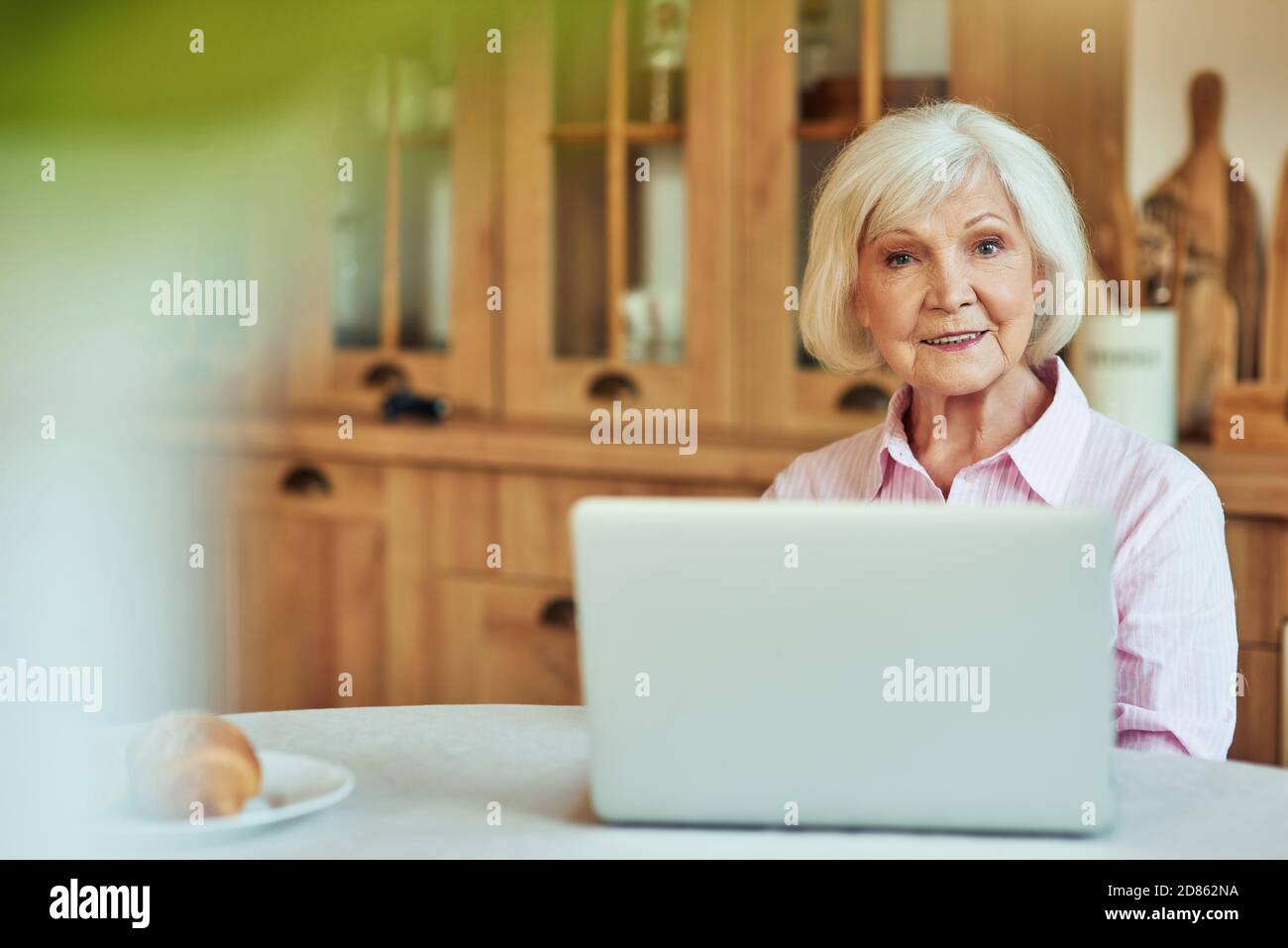 Die Hüfte nach oben lächelnde ältere Frau, die im Zimmer am Tisch sitzt, während sie mit einem Laptop arbeitet. Inländisches Lifestyle-Konzept Stockfoto