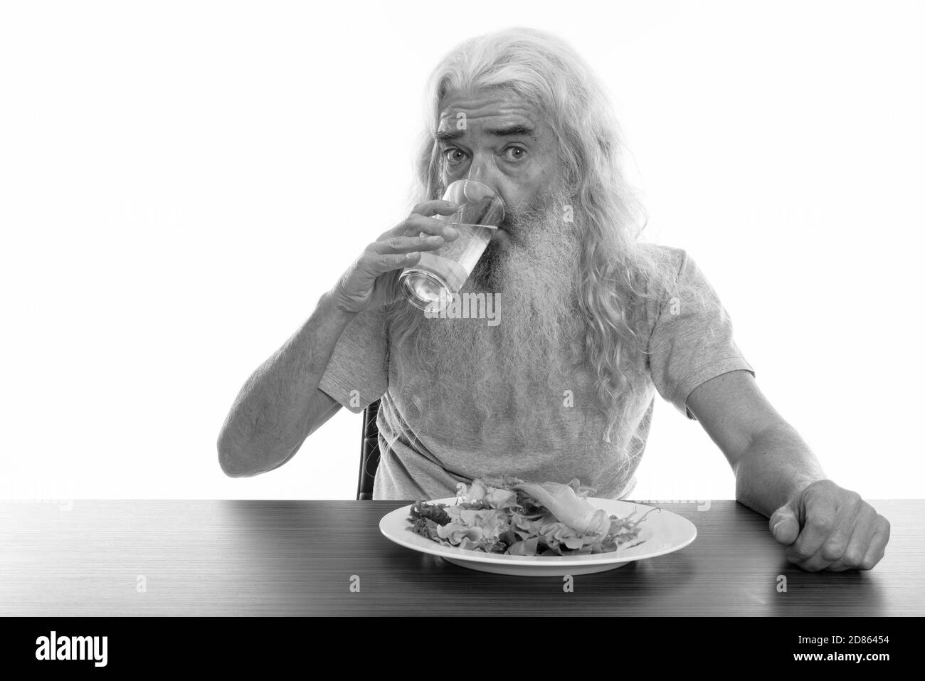 Studio shot der älteren bärtigen Mann trinken Glas Wasser mit Teller Salat auf hölzernen Tisch Stockfoto