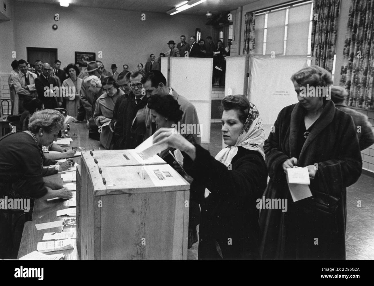 Die Wähler stehen am Wahltag an, um sich mit den Wahlbeamten zu erkundigen und Stimmzettel zu erhalten, auf denen sie ihre Stimmen abgeben können. Frau im Vordergrund hat ihre Stimmzettel markiert und trägt sie in die richtigen Boxen - eine für den Präsidentschaftskandidaten und eine für den Kongresskandidaten Dunn Loring, VA 11/8/1960. (Foto von Edwin Huffman/United States Information Agency/RBM Vintage Images) Stockfoto