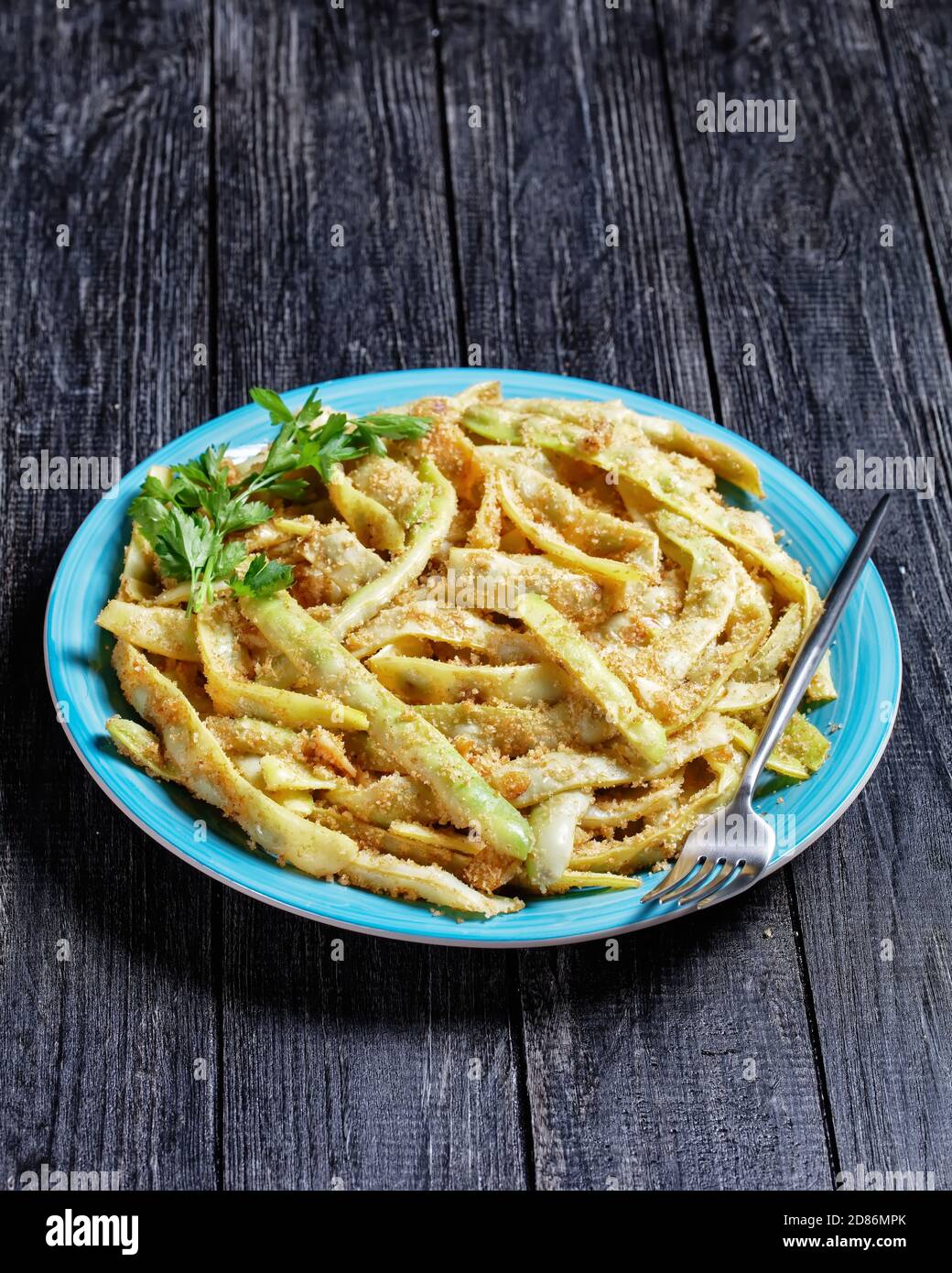 Goldene französische kalorienarme Wachsbohnen mit Knoblauch und Petersilie, mit Paniermehl überzogen und auf einer Pfanne gebraten, serviert auf einem blauen Teller mit Gabel auf einem dunklen Stockfoto