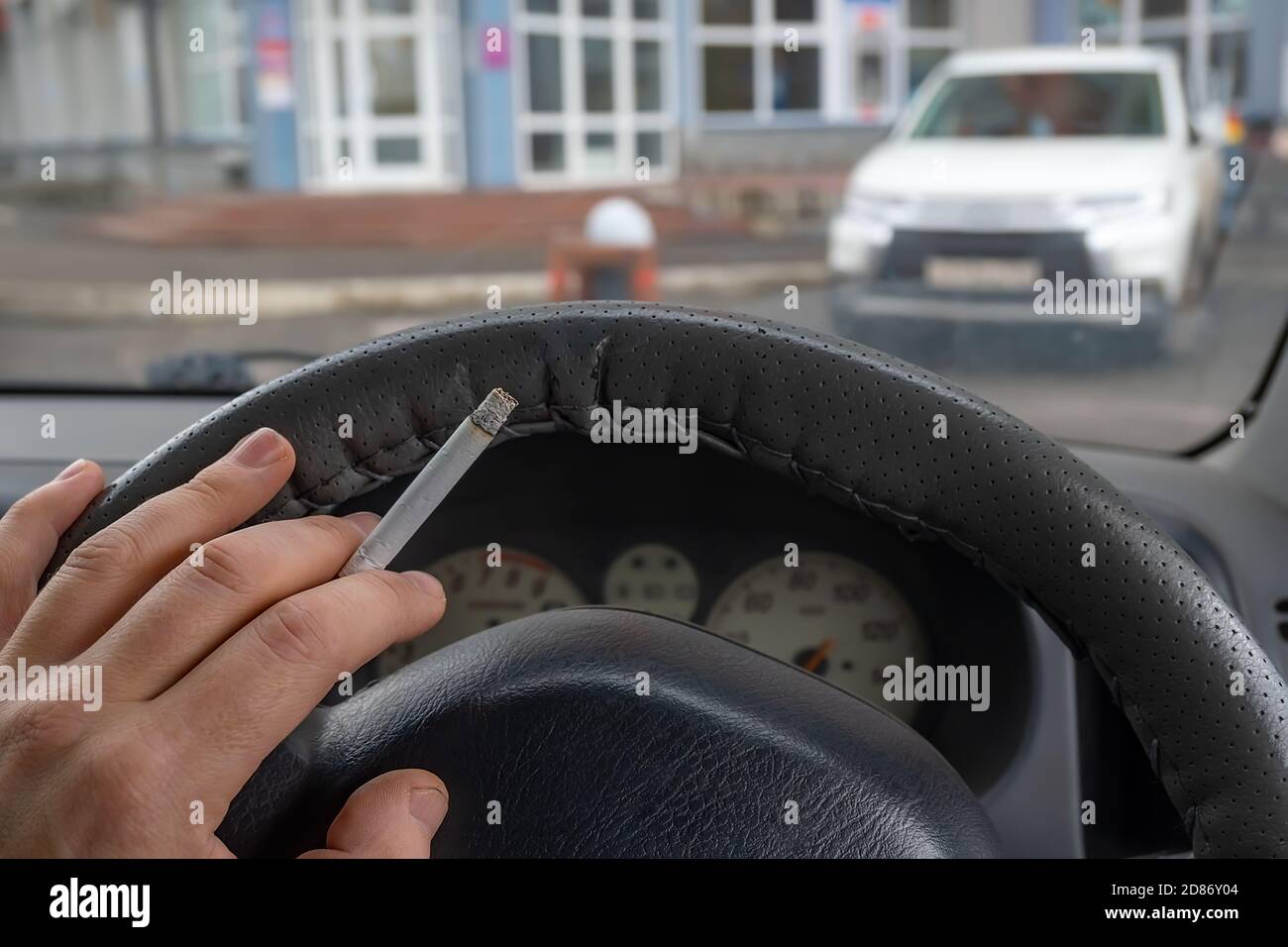 Fahrer Hand auf das Lenkrad des Autos, die eine Rauchen Zigarette hält, beobachten den Eingang zum Gebäude Stockfoto