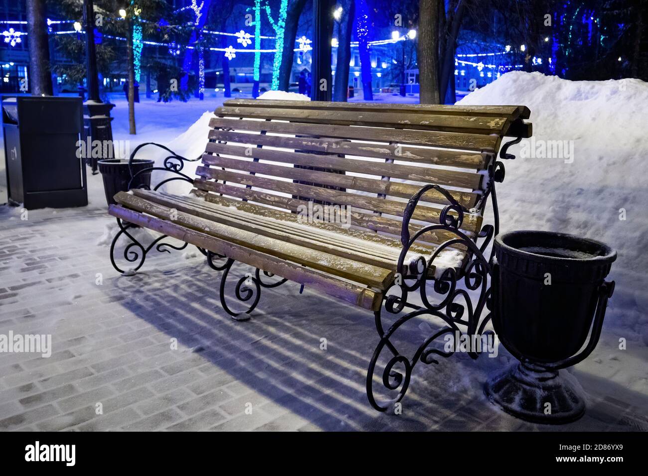 Leere braune Holzbank mit verzierten Mustern im Schnee unter Lampe Licht am Winterabend im Stadtpark Stockfoto