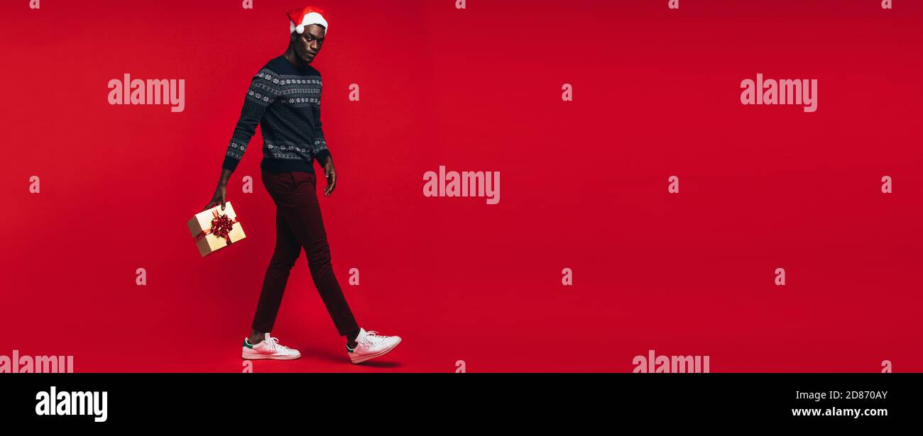 Junge afrikanische Mann, Santa hat springen mit Geschenkbox über den roten Hintergrund. Stattlichen schwarzen Kerl mit einem Weihnachtsgeschenk auf rotem Hintergrund. Stockfoto