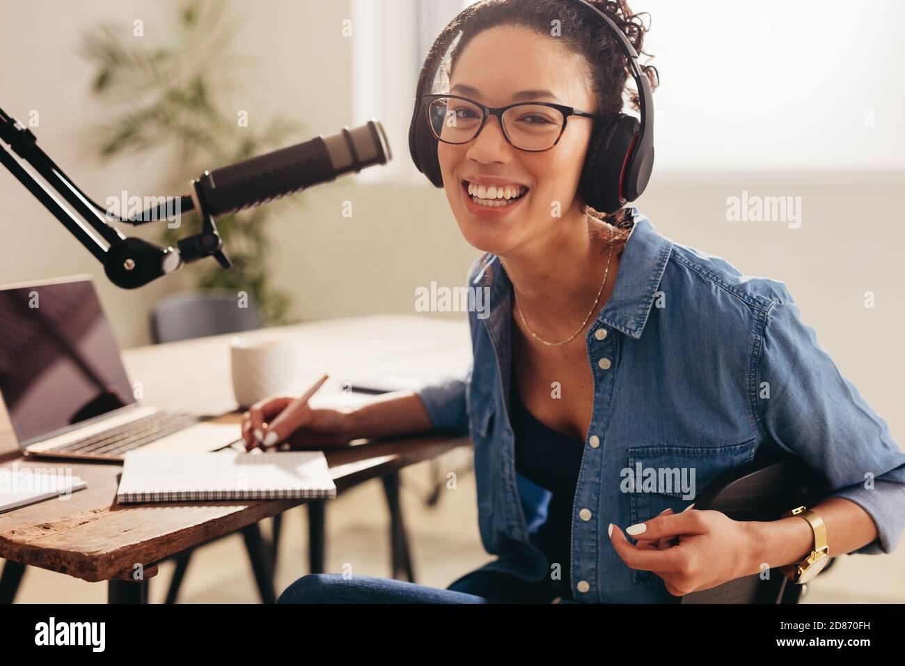 Fröhliche weibliche Podcast-Host arbeitet von zu Hause aus. Frau trägt Kopfhörer sitzen am Tisch mit Laptop, Mikrofon und einem Notizblock Blick auf Kamera und Stockfoto