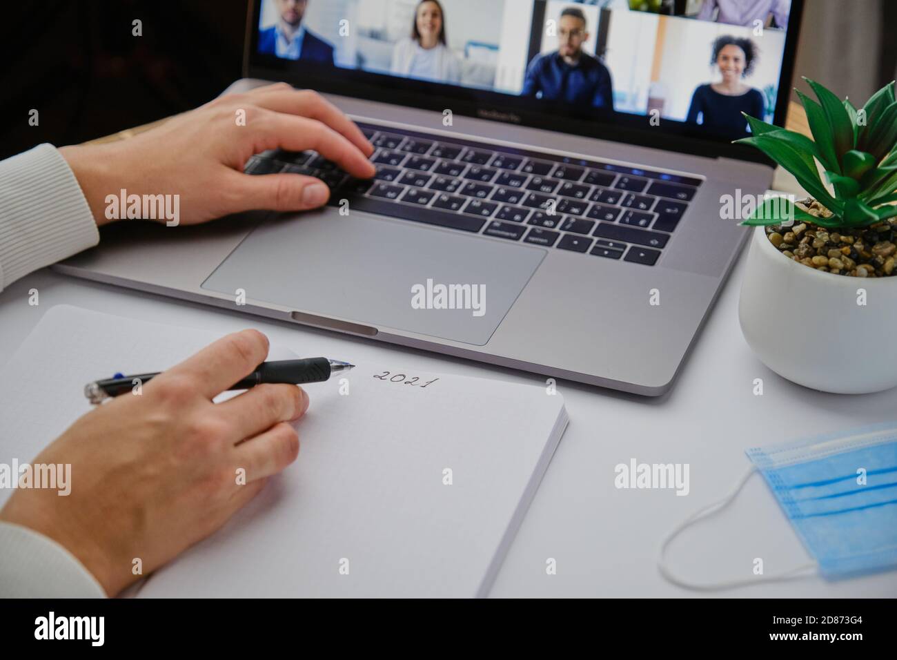 Home Office-Geschäftskonzept. Laptop auf einem weißen Schreibtisch mit Pflanzenmaske und Notizbuch. Frauenhände schreiben Stockfoto