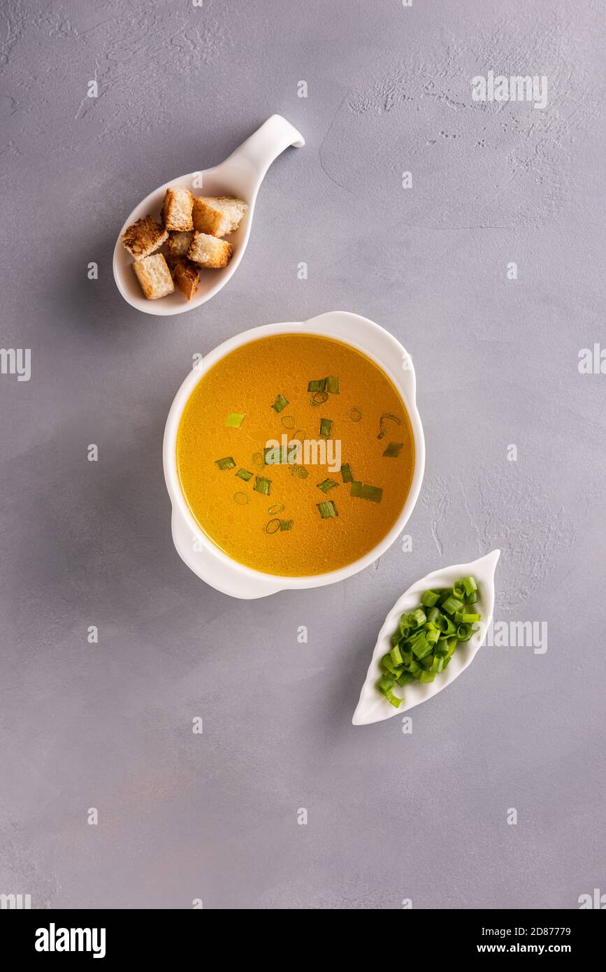 Bouillon in weißer Suppenschüssel, gehackte Frühlingszwiebeln oder Frühlingszwiebeln und Croutons auf grauem Steintisch Hintergrund. Direkt über der Ansicht. Vertikale Ausrichtung Stockfoto