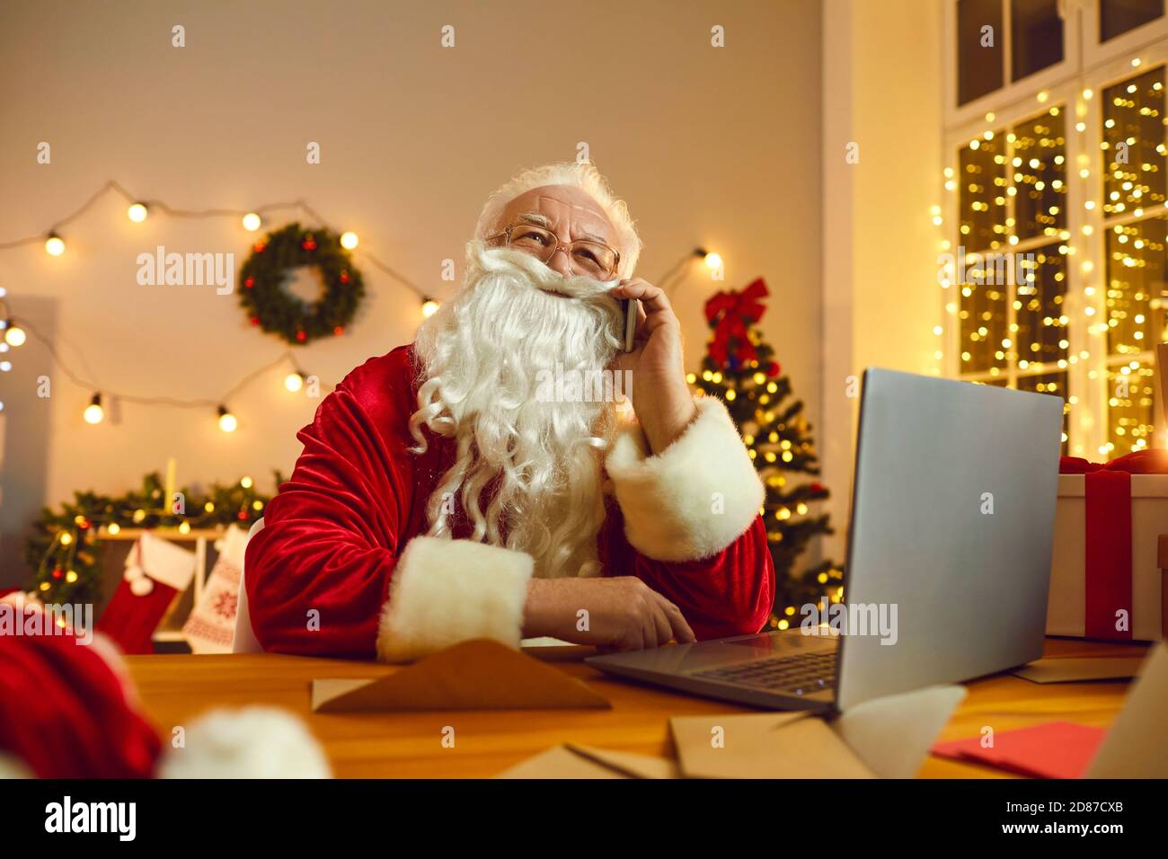 Der Weihnachtsmann arbeitet am Laptop, während er im Zimmer, das für Weihnachten dekoriert ist, telefoniert. Stockfoto